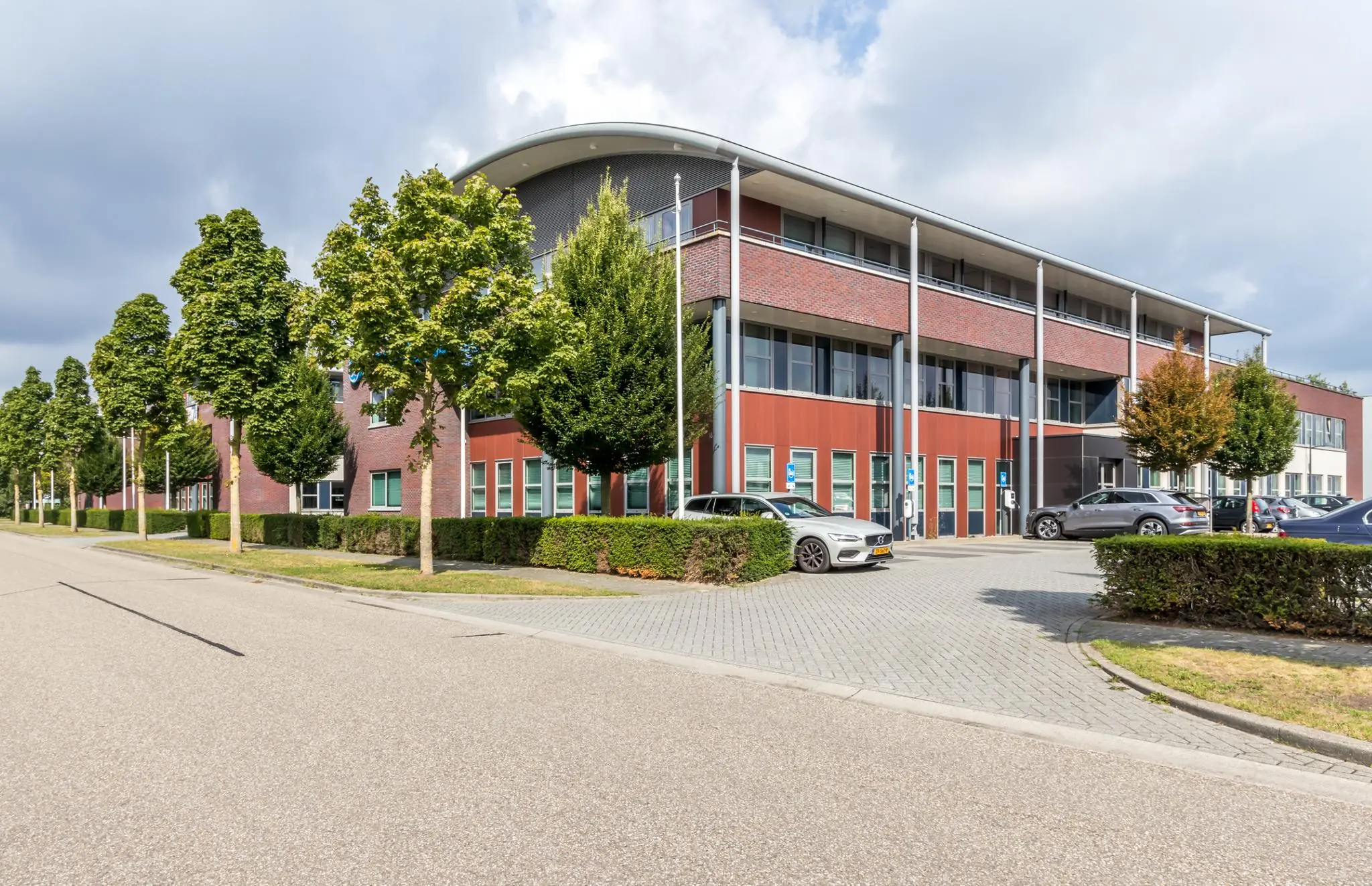 Modern kantoorgebouw aan De Wel met parkeerplaatsen en omliggende bomen.